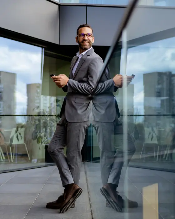 Smiling businessman in gray suit holding phone, leaning against glass wall in modern office building