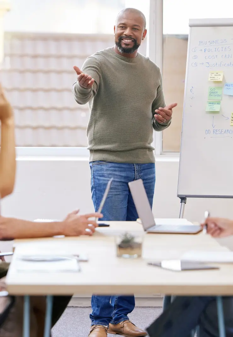 Man giving presentation during office meeting, standing by whiteboard with team seated around table
