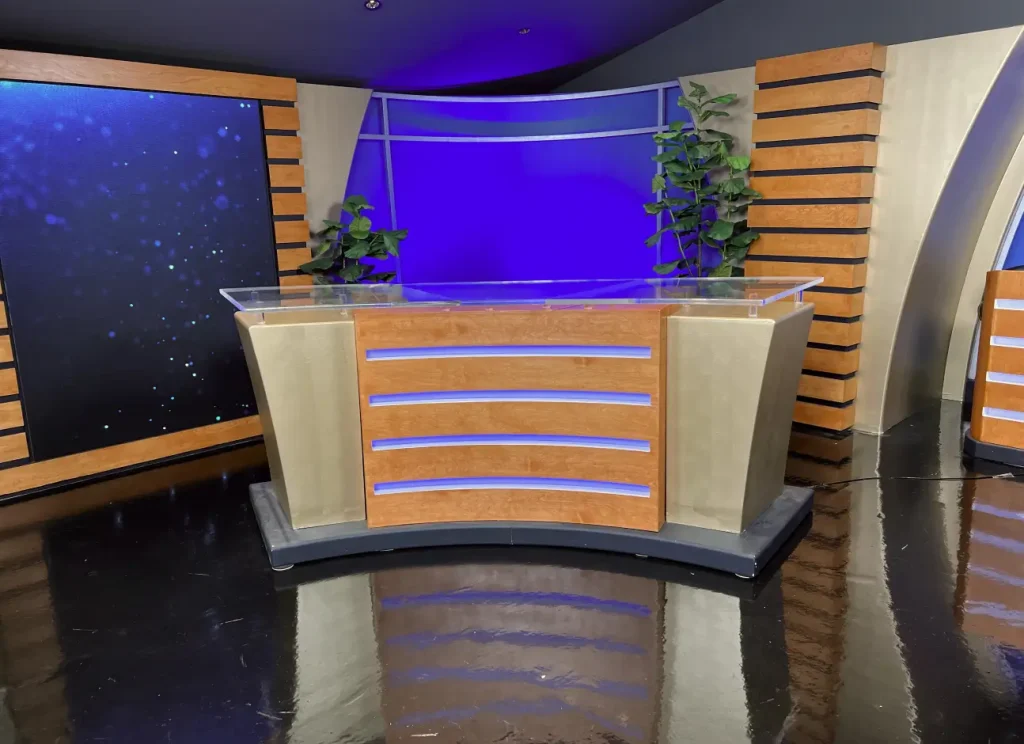 Modern news studio set featuring illuminated anchor desk, LED screen, blue backdrop, and decorative plants