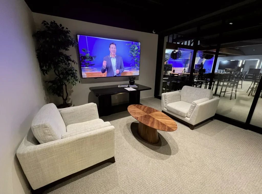 Modern office lounge with two chairs, wooden coffee table, wall-mounted TV, and glass wall view into studio