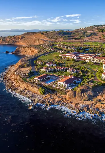 Aerial view of a luxury resort on a rocky coastline with ocean waves, green landscape, and villas by the sea